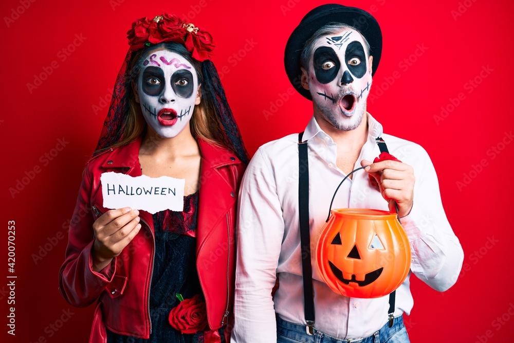 Couple wearing day of the dead costume holding pumpking and halloween paper afraid and shocked with surprise and amazed expression, fear and excited face.