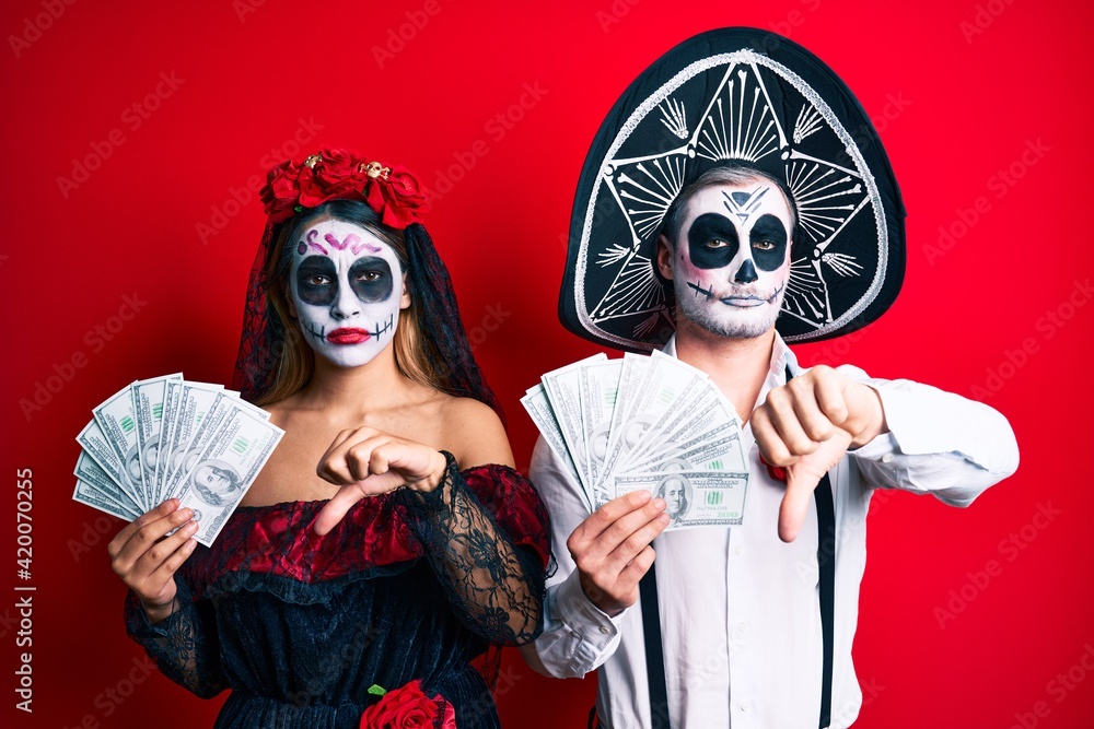 Couple wearing day of the dead costume holding dollars with angry face, negative sign showing dislike with thumbs down, rejection concept Couple wearing day of the dead costume holding dollars with angry face, negative sign showing dislike with thumbs down, rejection concept