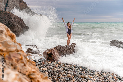 A beautiful girl in a white shirt and black swimsuit stands on a rock  big waves with white foam. A cloudy stormy day at sea  with clouds and big waves hitting the rocks.