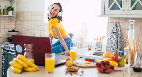 Excited beautiful happy young woman in headphones is dancing and cooking in the kitchen at home