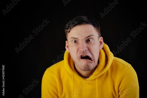 Portrait of young man in yellow hoodie with grimacing face. Close up of funny guy monkeying on black background.