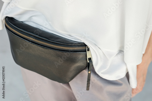 Close-up. A woman in a white blouse and gray pants wears a black leather banana bag. Hip Pouch For Women