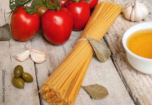 Raw spaghetti pasta next to fresh red tomatoes on a branch, garlic, olive oil and bay leaves on a wooden surface.