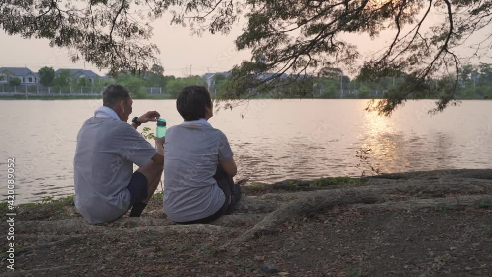 happy senior couple sitting near lake view offer bottle of water in evening sunset. smiley sweet asian elderly lover romantic sharing good moment together. sport mature in sportswear resting at park.