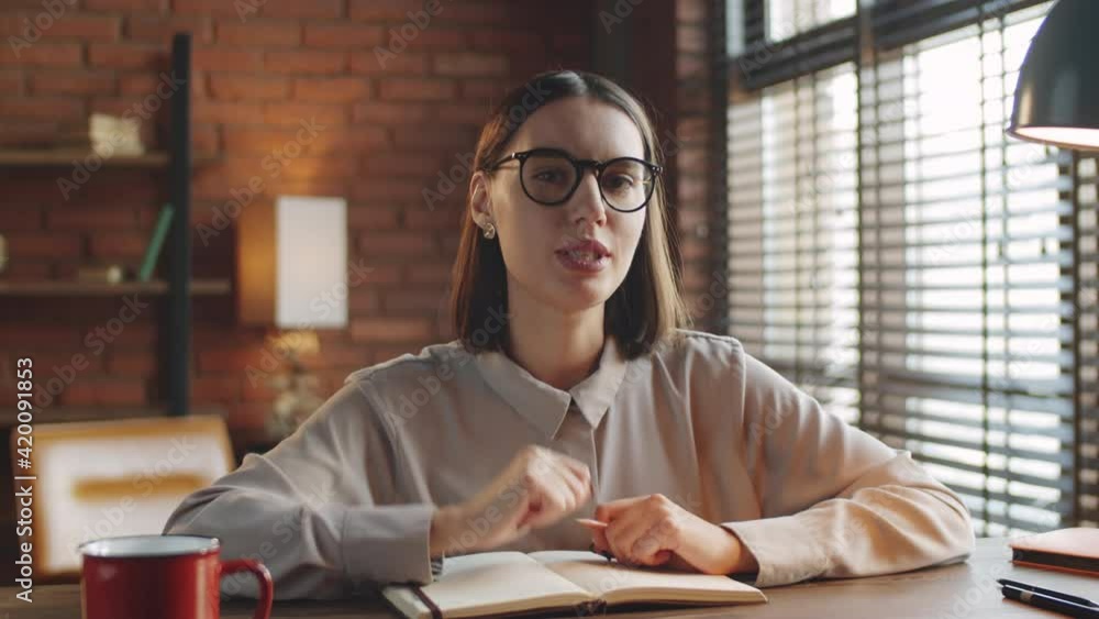 Young beautiful woman sitting at desk in loft office, taking notes, smiling and speaking at camera via online video call Young beautiful woman sitting at desk in loft office, taking notes, smiling and speaking at camera via online video call