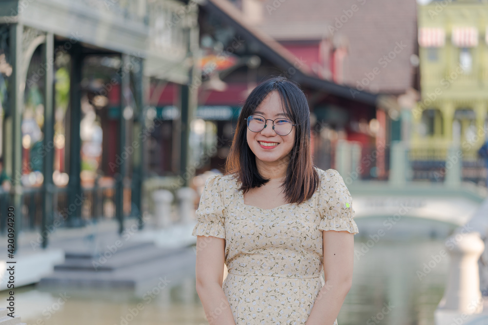 portrait of Asian woman with eyeglasses smiling with happiness near river in colorful town, woman in casual clothes relaxing on bridge