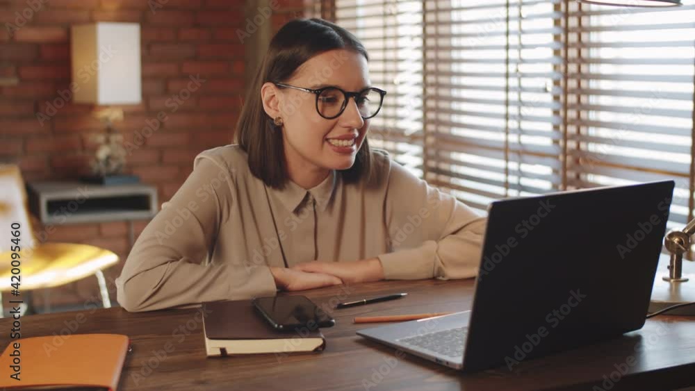 Young beautiful businesswoman sitting at desk in loft office, smiling and speaking on online video call on laptop Young beautiful businesswoman sitting at desk in loft office, smiling and speaking on online video call on laptop