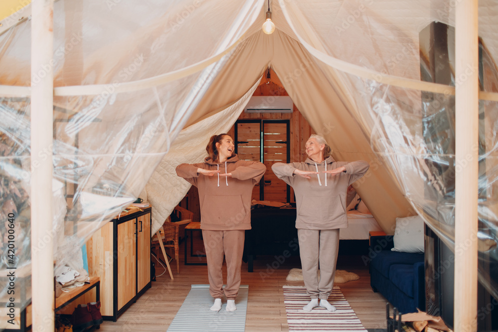 Family doing exercises sports indoor. Young and senior elderly woman relaxing at glamping camping tent. Mother and daughter modern at fitness vacation lifestyle concept. - obrazy, fototapety, plakaty 