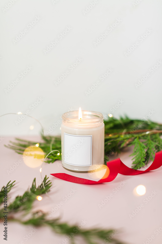 Blank burning candle surrounded by pine branches, red ribbon, and lights on pink background with copy space Blank burning candle surrounded by pine branches, red ribbon, and lights on pink background with copy space