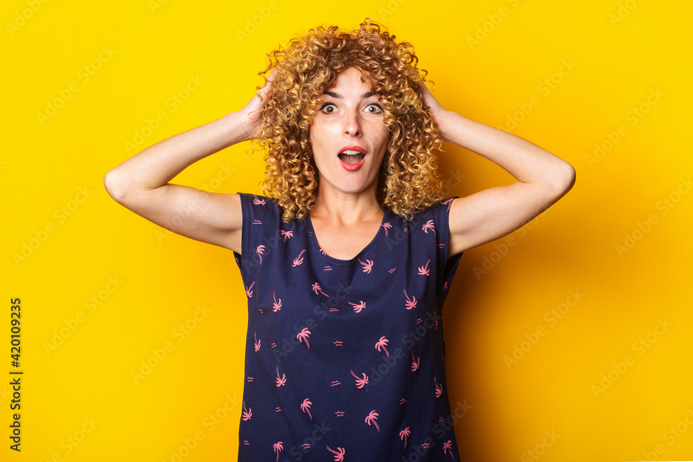 shocked surprised beautiful curly young woman holding her head with her hands on a yellow background. shocked surprised beautiful curly young woman holding her head with her hands on a yellow background.