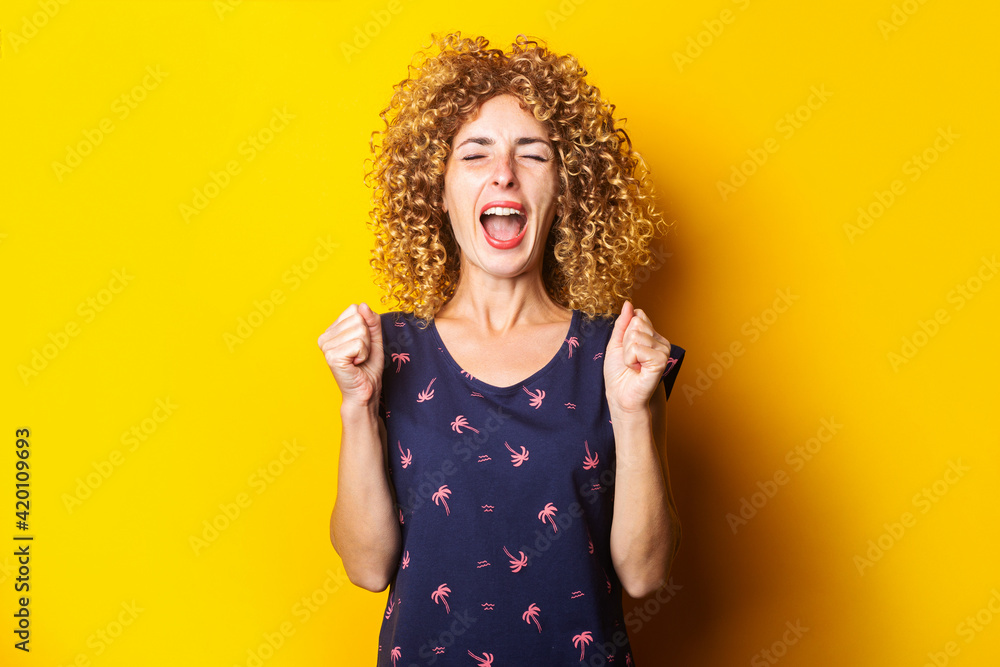 curly young woman screaming with closed clenched fists on yellow background. curly young woman screaming with closed clenched fists on yellow background.