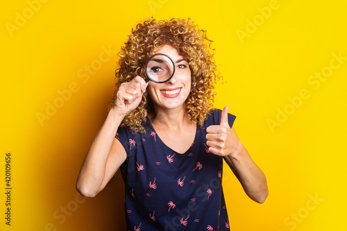 curly young woman looking at camera through magnifying glass showing thumbs up gesture on yellow background.