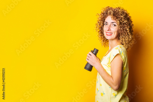 smiling curly girl holding binoculars in her hands on a yellow background.