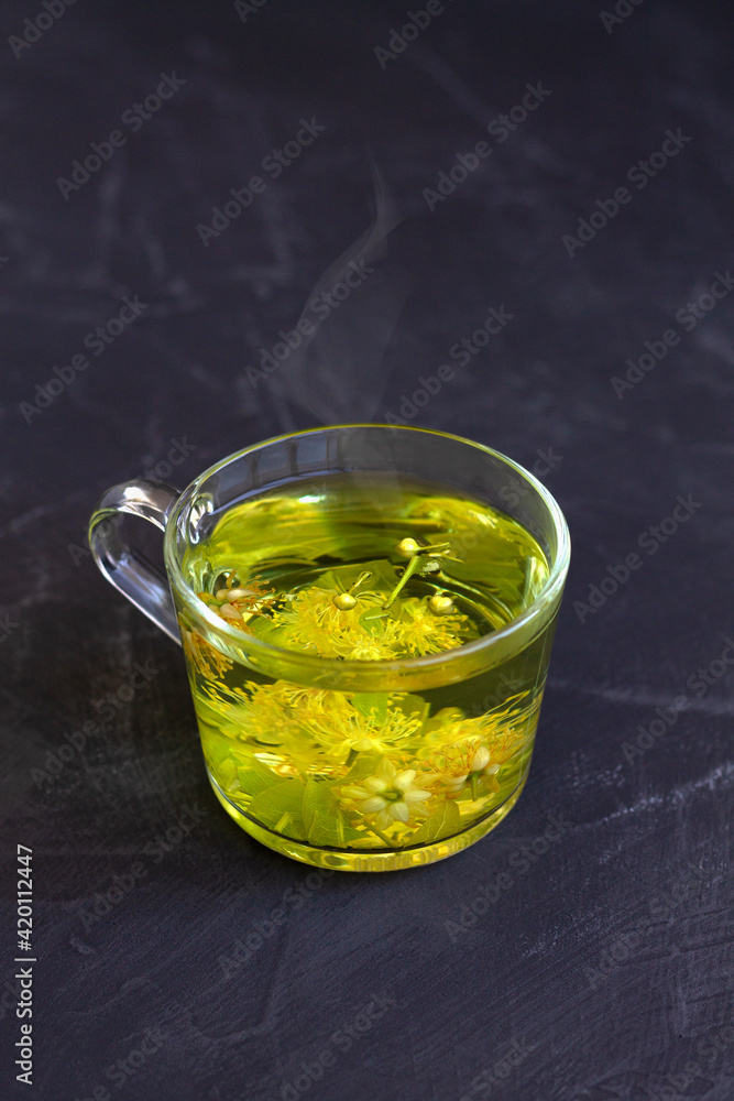 hot linden flowers tea.Cup of hot herbal tea with linden fresh flowers on a black table.Healthy lifestyle.glass cup of tea with green leaves on a black background.hot drinks.copy space.vertical hot linden flowers tea.Cup of hot herbal tea with linden fresh flowers on a black table.Healthy lifestyle.glass cup of tea with green leaves on a black background.hot drinks.copy space.vertical