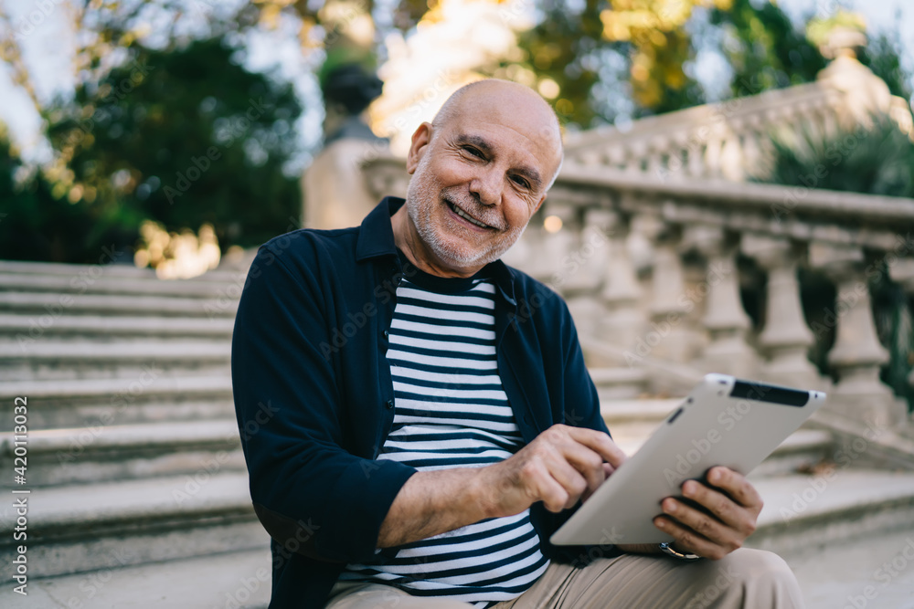 Half length portrait of happy senior traveller with modern touch pad gadget smiling at camera during free time in city, elderly male tourist 60s with digital tablet posing during vacations trip