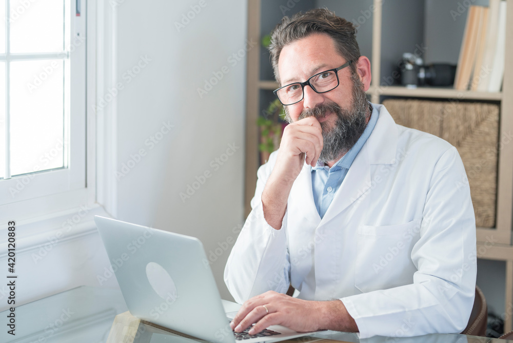 Smiling old male doctor in glasses and white uniform sit at desk in hospital work on laptop write in journal, happy mature senior man physician fill patient medical history anamnesis on computer