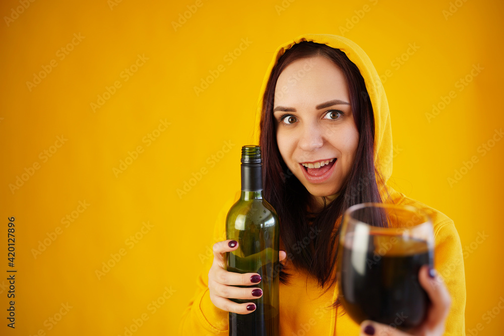 Relaxed young woman poses with bottle and glass of red wine on yellow background. Adult happy brunette holds alcohol in hands. Relaxed young woman poses with bottle and glass of red wine on yellow background. Adult happy brunette holds alcohol in hands.