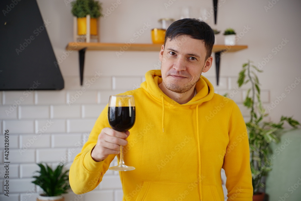 Relaxed young man poses with glass of red wine, standing at kitchen table. Adult guy resting with alcohol in kitchen. Relaxed young man poses with glass of red wine, standing at kitchen table. Adult guy resting with alcohol in kitchen.