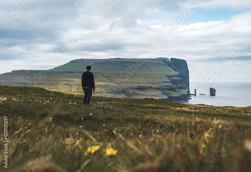 Young man tourist standing in front of Risin and Kellingin rocks in the sea as seen from Tijornuvik bay on Streymoy on the Faroe Islands, Denmark, Europe