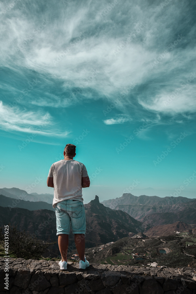 Naklejka premium Young man standing on top of the mountain on the edge. Wearing beige t-shirt, denim shorts and white shoes. Mountain landscape. 