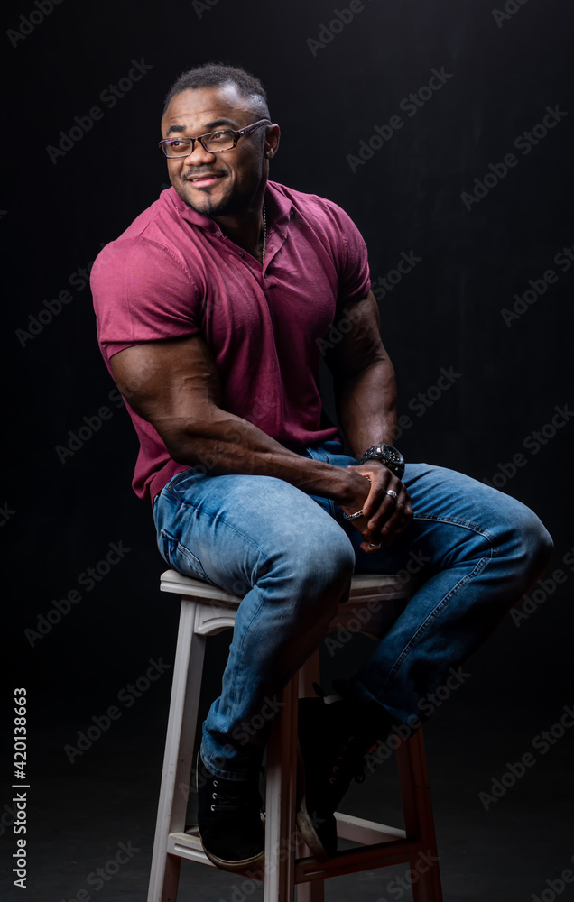 Young handsome man in red shirt and jeans sitting on chair. Athletic muscular male posing for camera on chair. Young handsome man in red shirt and jeans sitting on chair. Athletic muscular male posing for camera on chair.