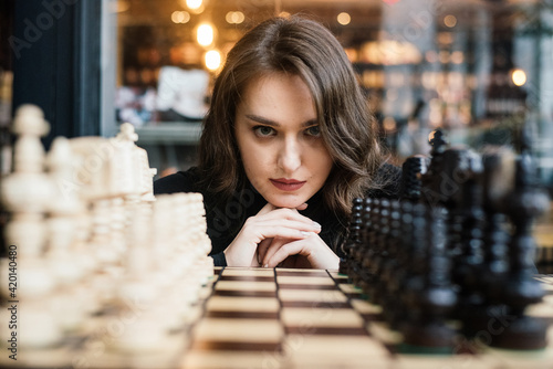 Beautiful girl prepared to play chess, plays Queen's Gambit, smart and pensive face. White and black chess pieces are displayed on board. Mental game, long time to last, not for fools.