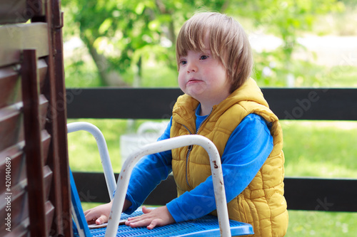 Portrait of cute adorable blondy toddler disabled boy with down sydrome while standing and holding chair. Dressed with yellow vest and blue sweatshirt