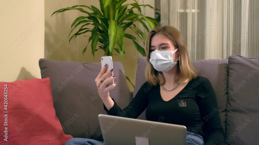 Young Turkish woman with a facial mask, having a video call on her smartphone while sitting on the sofa. There is a laptop computer on her lap, coffee cup and a hand sanitizer bottole on the table. Young Turkish woman with a facial mask, having a video call on her smartphone while sitting on the sofa. There is a laptop computer on her lap, coffee cup and a hand sanitizer bottole on the table.