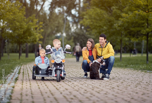 young caucasian parents playing in park with their sons in motorcycle toy and a dog