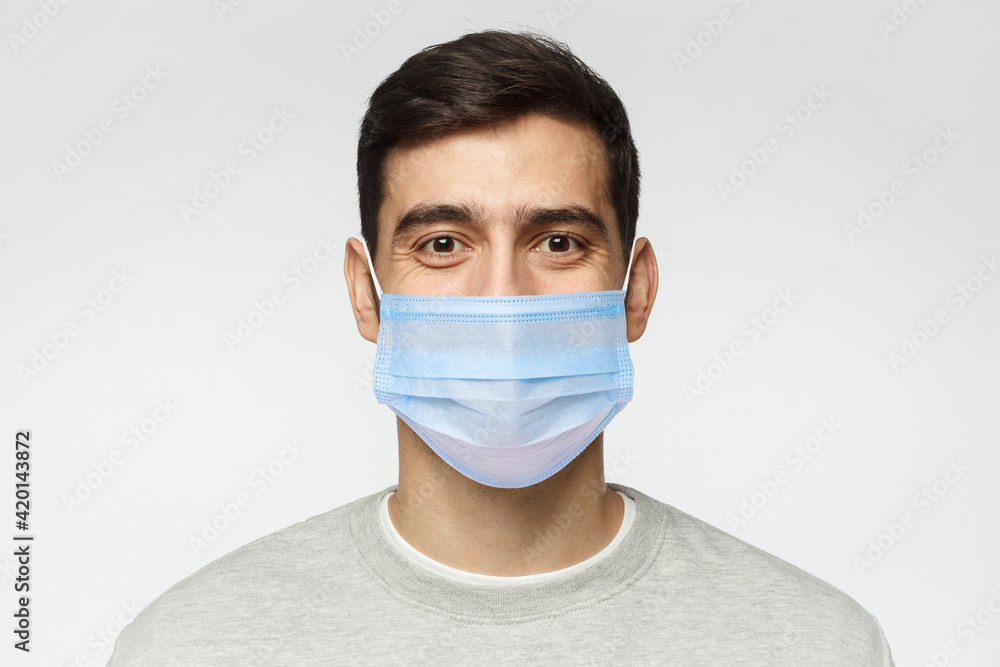 Young handsome man wearing sweatshirt and medical mask, standing isolated on gray background Young handsome man wearing sweatshirt and medical mask, standing isolated on gray background