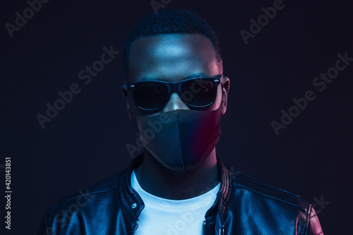 African male in sunglasses and coronavirus mask standing on night street in neon light
