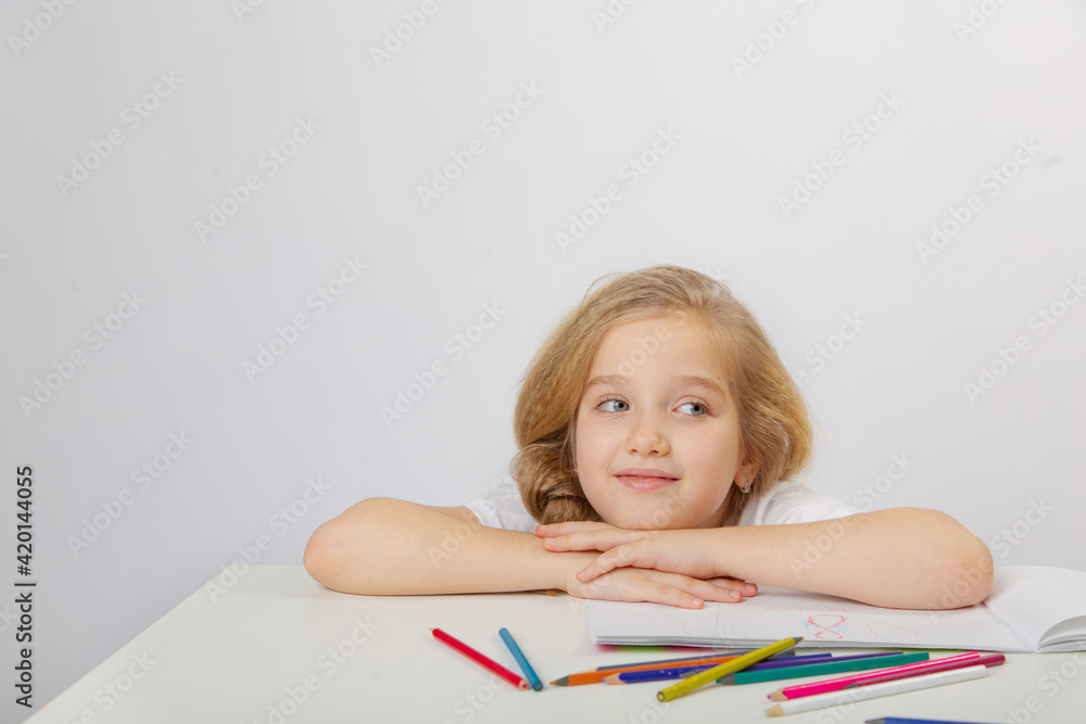A small cute schoolgirl girl with blonde hair draws an inscription in an album with colored pencils on a light background A small cute schoolgirl girl with blonde hair draws an inscription in an album with colored pencils on a light background