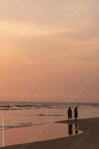Kobiety spacerujące brzegiem oceanu o zachodzie słońca, ciemno pomarańczowy nastrojowy krajobraz.
