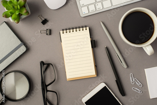 Overhead photo of notebook keyboard computer mouse magnifier pens phone plant glasses cup of coffee and paperclips isolated on the grey background