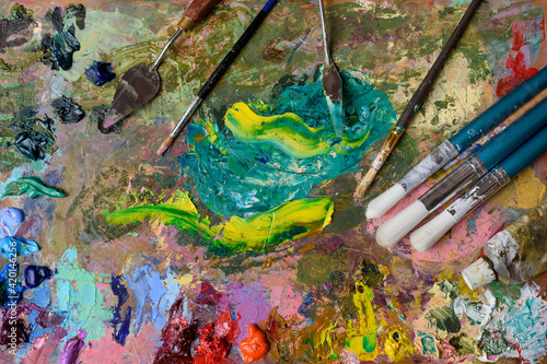 The texture of the artist's wooden palette with brushes, paints and palette knives. Mixed bright oil paints of different colors and saturations. Close-up art objects and tools for creativity.