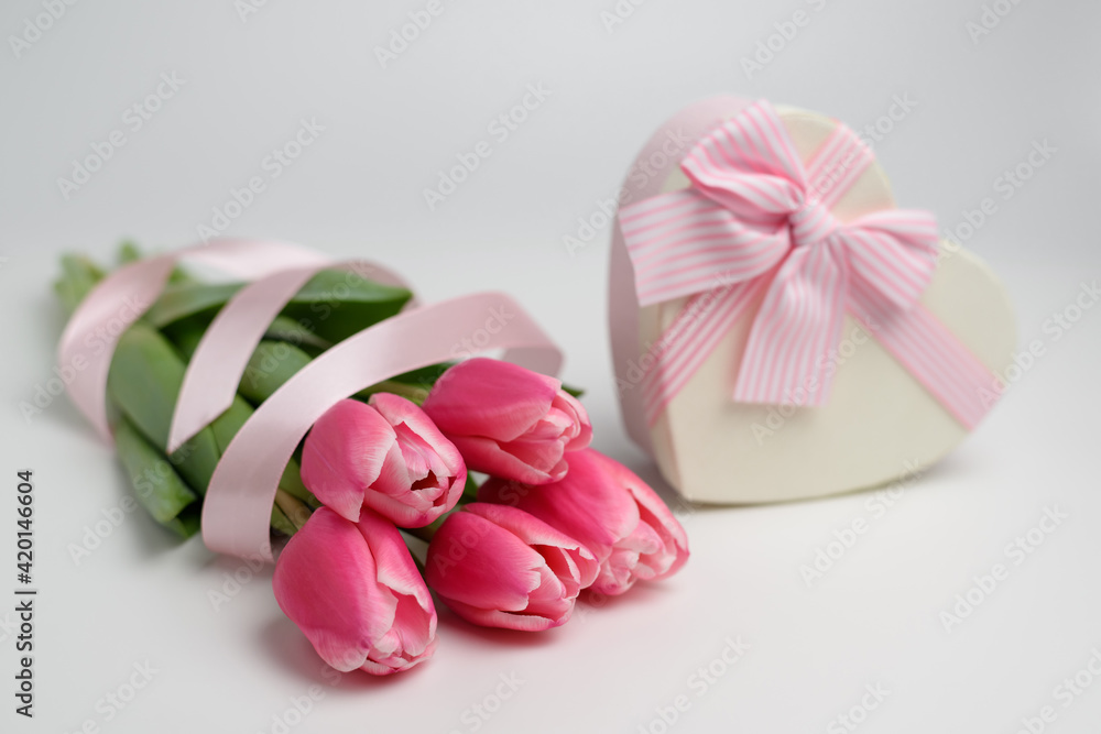 Beautiful tulips of pink color on a white background near a gift box in the shape of a heart with a ribbon and a beautifully tied pink bow. The concept of a gift and surprise for a loved one. Beautiful tulips of pink color on a white background near a gift box in the shape of a heart with a ribbon and a beautifully tied pink bow. The concept of a gift and surprise for a loved one.
