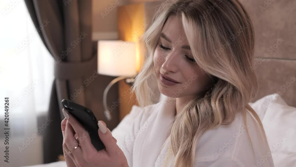 Young female model pretty lady woman wearing textile bath robe sitting on white bed blanket in modern hotel suite using her smartphone typing message searching information online chatting with friend. Young female model pretty lady woman wearing textile bath robe sitting on white bed blanket in modern hotel suite using her smartphone typing message searching information online chatting with friend.