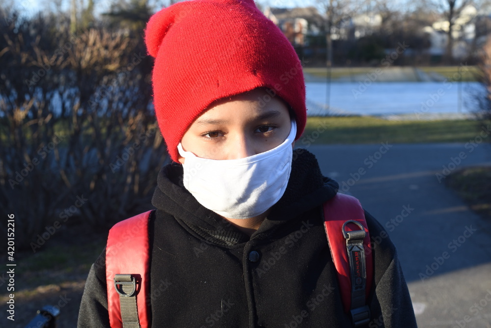 Cool mixed boy in mask and in red hat and with red backpack going to school after pandemic time. Schoolkids in masks.