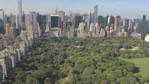 AERIAL ESTABLISHING SHOT: Flying above Central Park along 5th Avenue and towards Downtown Manhattan in sunny New York City. Tall glassy skyscrapers and condominium buildings overlooking Central Park