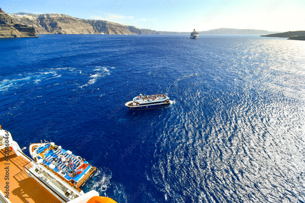 A tender boat pulls alongside a massive cruise ship with another boat and ship in the distance inside the caldera of Santorini Greece