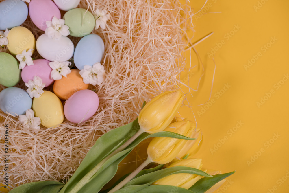 multicolored easter eggs on hay and near yellow tulips on yellow background multicolored easter eggs on hay and near yellow tulips on yellow background