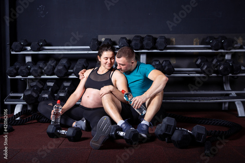 Beautiful sportive pregnant couple sitting on the floor hugging in the gym after a workout smiling and rejoicing. Fitness and healthy lifestyle during pregnancy. Family healthy lifestyle concept