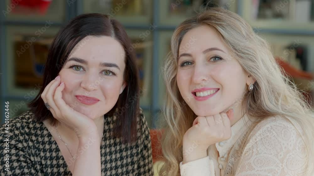 Two positive young women with toothy smile looking at each other and posing. Portrait of happy carefree relaxed Caucasian female friends in cafe or restaurant indoors. Lifestyle and beauty concept. Two positive young women with toothy smile looking at each other and posing. Portrait of happy carefree relaxed Caucasian female friends in cafe or restaurant indoors. Lifestyle and beauty concept.