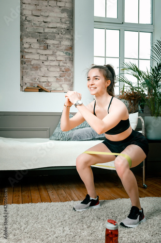 A strong and beautiful sports fitness girl in sportswear doing squats with sports fitness bands in her bright and airy bedroom with a minimalistic interior.
