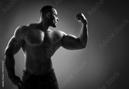Black and white portrait of brutal strong man athlete in jeans and half naked shirtless standing showing strong huge pumped up biceps over grey background. Sport men body concept © Dmitry Lobanov