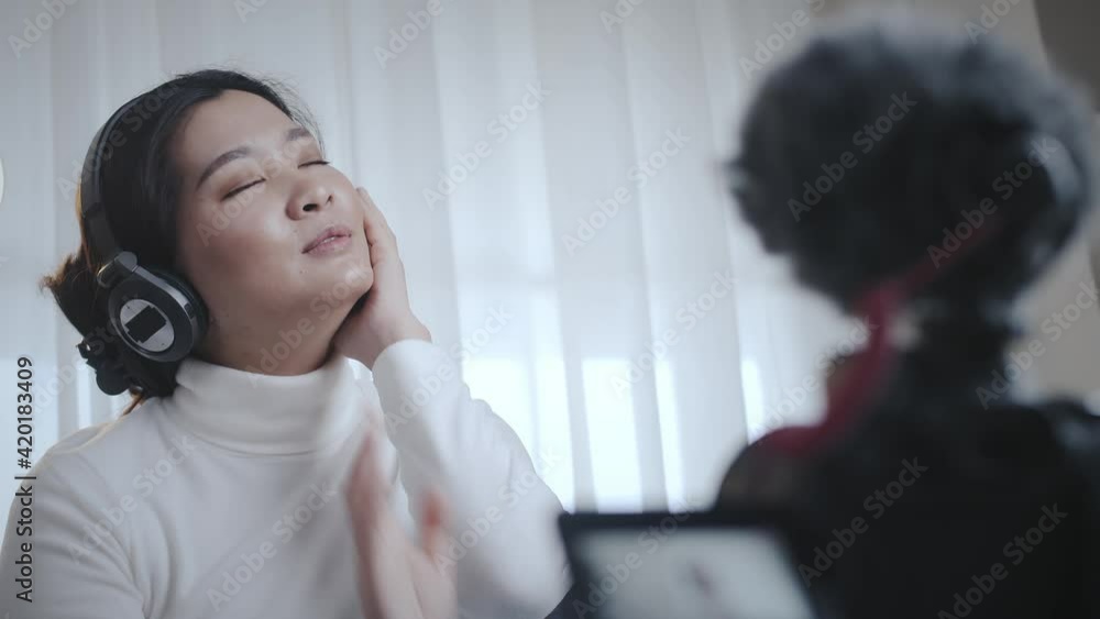 Happy cheerful Asian woman wearing wireless headphones recording video for social media sitting on the sofa at home lifestyle and pleasure Happy cheerful Asian woman wearing wireless headphones recording video for social media sitting on the sofa at home lifestyle and pleasure