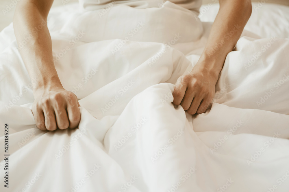 A man sitting on the bed and holding a white blanket on his hand. Bedroom with white bed and sunlight. A man sitting on the bed and holding a white blanket on his hand. Bedroom with white bed and sunlight.