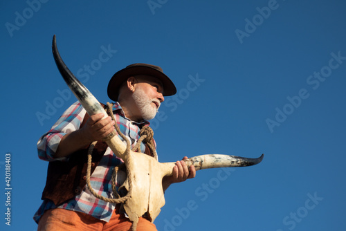 Old western cowboy with buffalo bone or cow skull. Senior bearded man with brown jacket and hat at wild west.