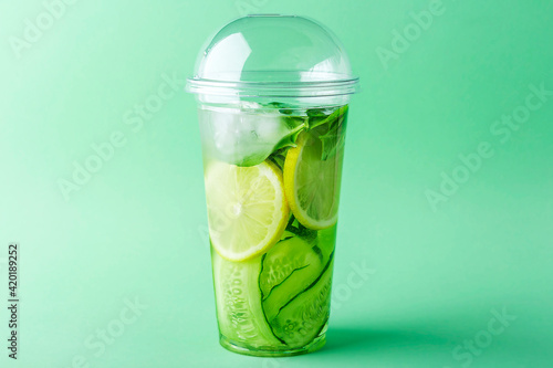 Fresh cool detox drink with cucumber, lemon and basil in plastic cup on green background. Tasty infused water to go. Proper nutrition and healthy eating. Fitness diet. Copy space for text, for menu.