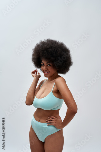 Playful curvy young female model with afro hair style wearing blue underwear smiling at camera, posing isolated over light background